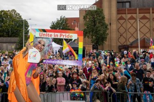 A large crowd celebrating Pride, with a performer in drag holding a colorful sign that says 'Unite in Pride' and '#UniteinPride', surrounded by people wearing rainbow attire and accessories.