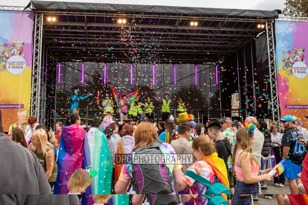 A vibrant scene at a pride festival featuring a stage adorned with colorful decorations. Confetti is falling over a crowd of diverse attendees, many wearing pride flags and colorful outfits. Some people are dancing while others are enjoying the festivities.