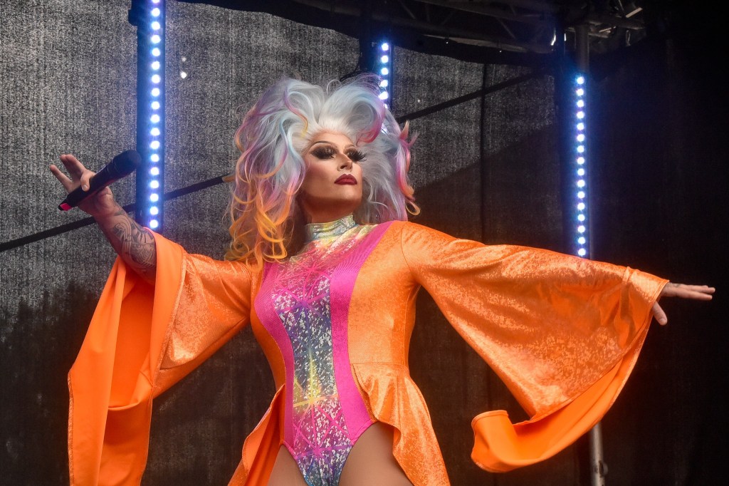 A performer with colorful hair and an orange outfit, singing on stage with a microphone, illuminated by stage lights.