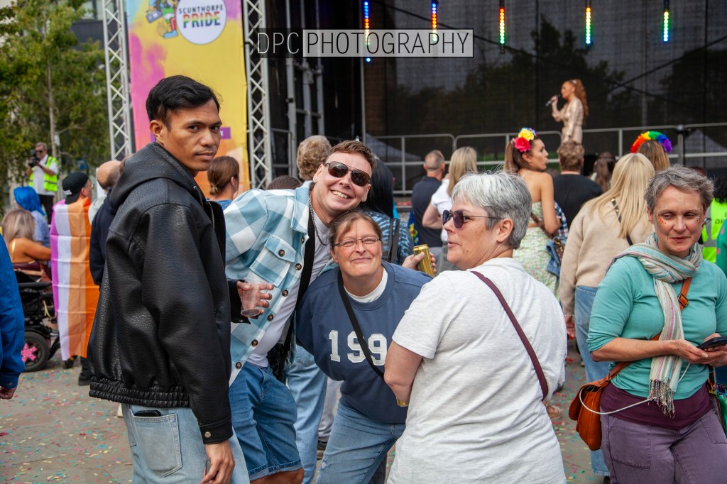 A group of four people smiling and posing for the camera at a lively outdoor event. They are surrounded by a crowd and colorful decorations in the background, with a performer visible on stage.