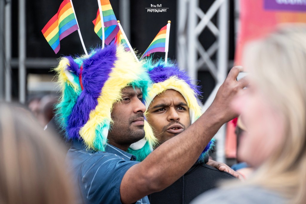 Two men wearing colorful furry hats with rainbow flags are taking a selfie at a celebration event.