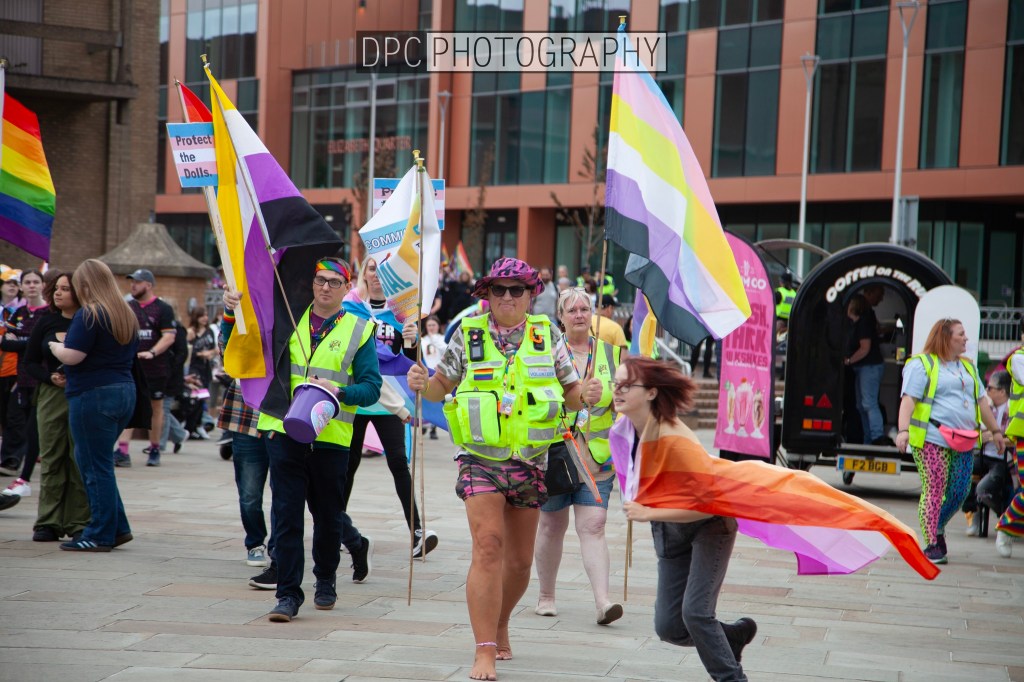 A group of people marching in a pride parade, holding colorful flags including a rainbow flag and a transgender flag, with some participants wearing vibrant clothing.