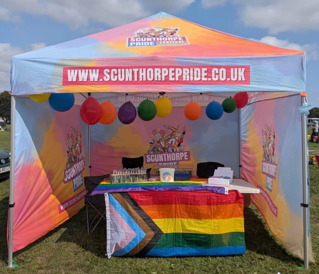 A colorful Scunthorpe Pride festival tent decorated with balloons and a rainbow flag, displaying promotional materials and donation information.