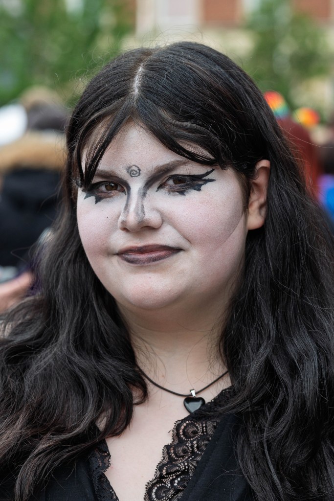 Close-up portrait of a young person with elaborate face makeup featuring dark eyeliner and lipstick, wearing a black outfit and a heart-shaped necklace.