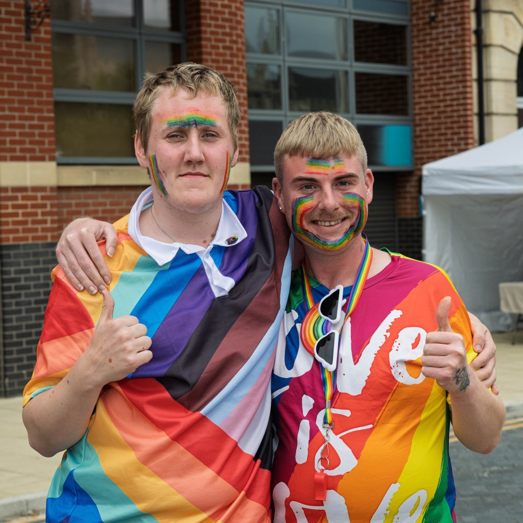 Two men smiling and posing together, both wearing colorful rainbow-themed shirts, celebrating pride with face paint and accessories.