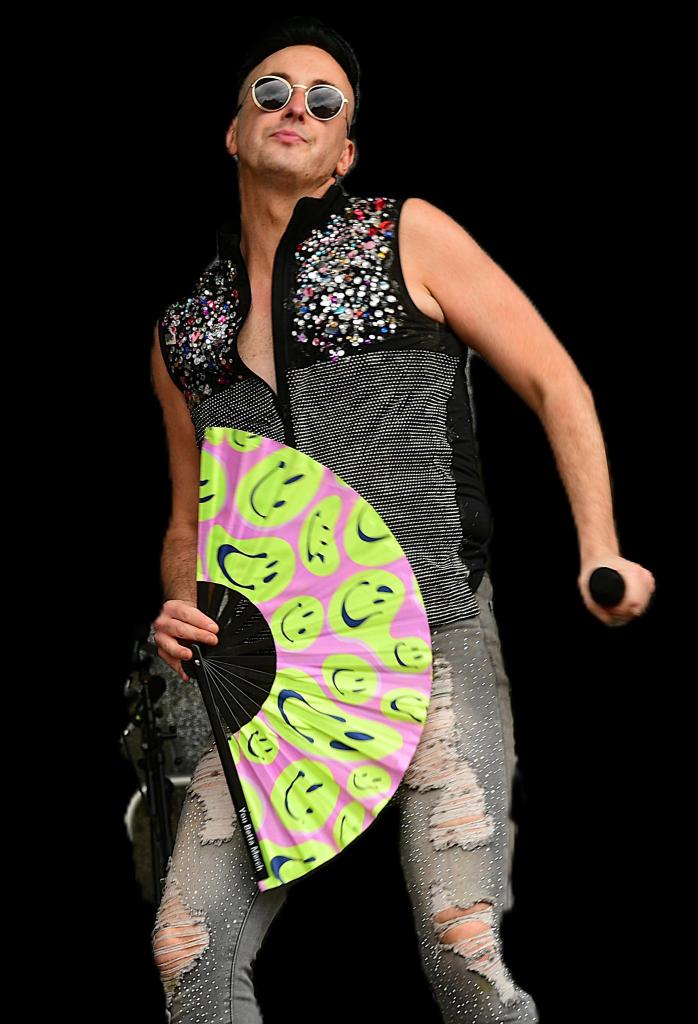A performer wearing sunglasses and a bedazzled black sleeveless top holds a colorful fan decorated with smiley faces while striking a pose on stage.