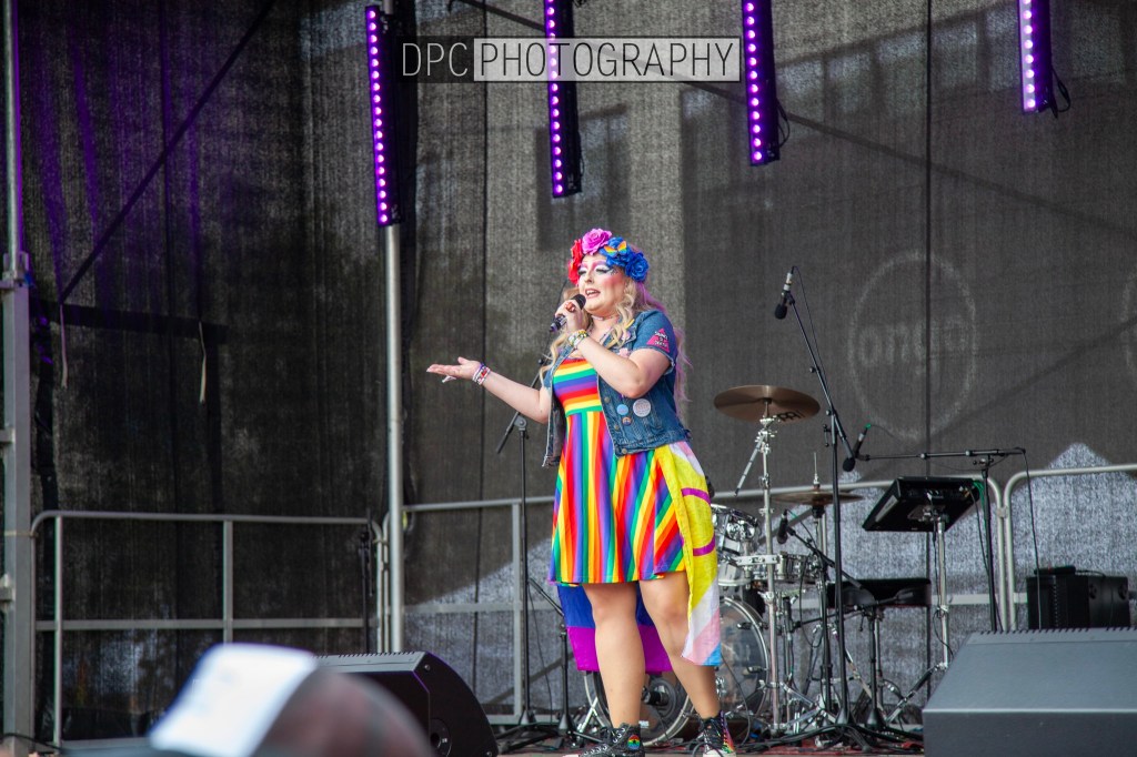 A performer on stage wearing a colorful rainbow dress and floral headpiece, singing into a microphone with a drum set in the background.