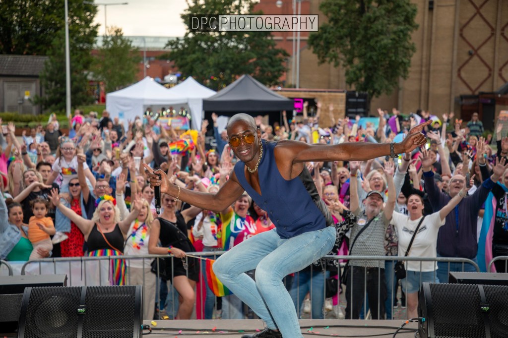 A performer energetically engaging with a lively crowd at an outdoor event, with people cheering and waving in the background.