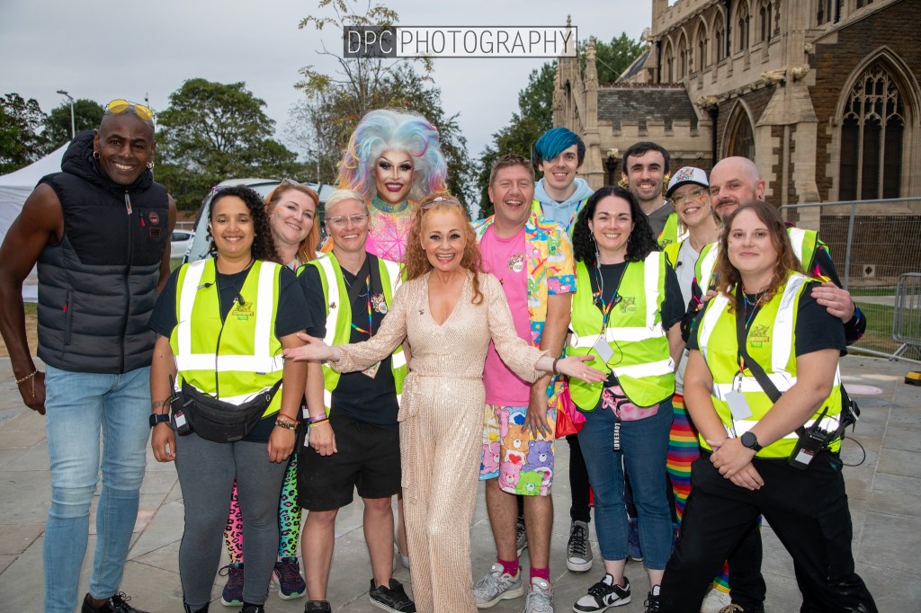 A group photo featuring a diverse team of individuals, including a performer in a colorful outfit and dramatic makeup, smiling together outdoors. Most attendees are wearing high-visibility jackets, conveying a sense of teamwork and celebration.