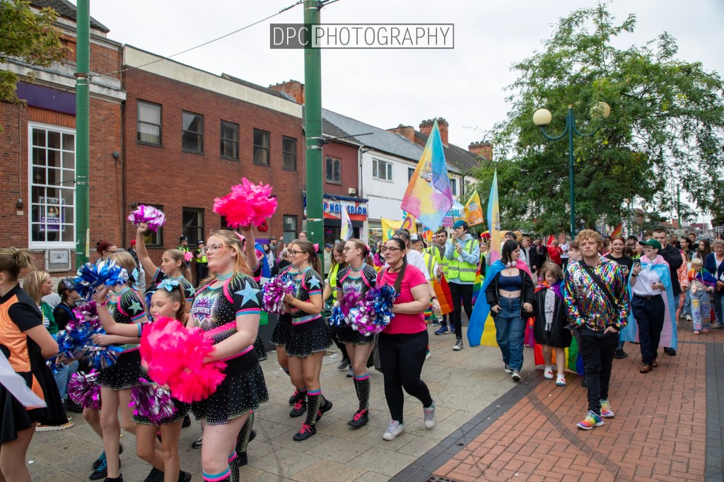 A vibrant parade featuring participants dressed in colorful outfits, dancing and holding pom-poms, celebrating diversity and inclusion in a lively street setting.