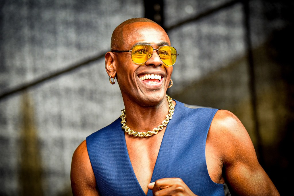 A smiling individual wearing yellow sunglasses and a blue sleeveless top, adorned with a chain necklace, poses confidently, showcasing their upbeat personality.