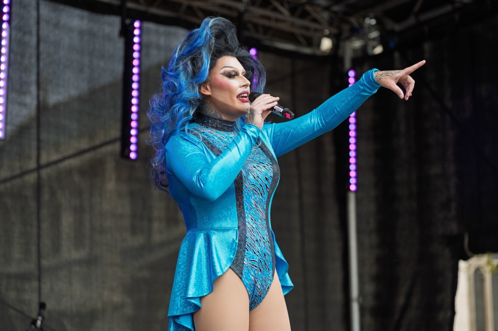 A performer in a blue outfit with dramatic hair gestures while singing into a microphone on stage.