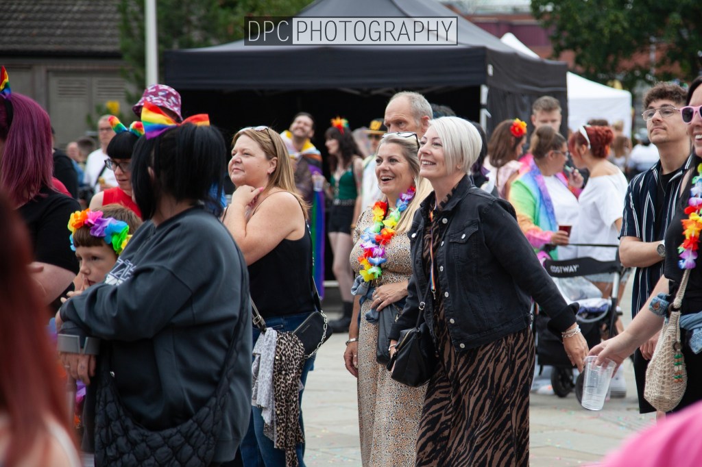A diverse group of people enjoying a lively event, wearing colorful attire and smiling, with decorations and a festive atmosphere in the background.
