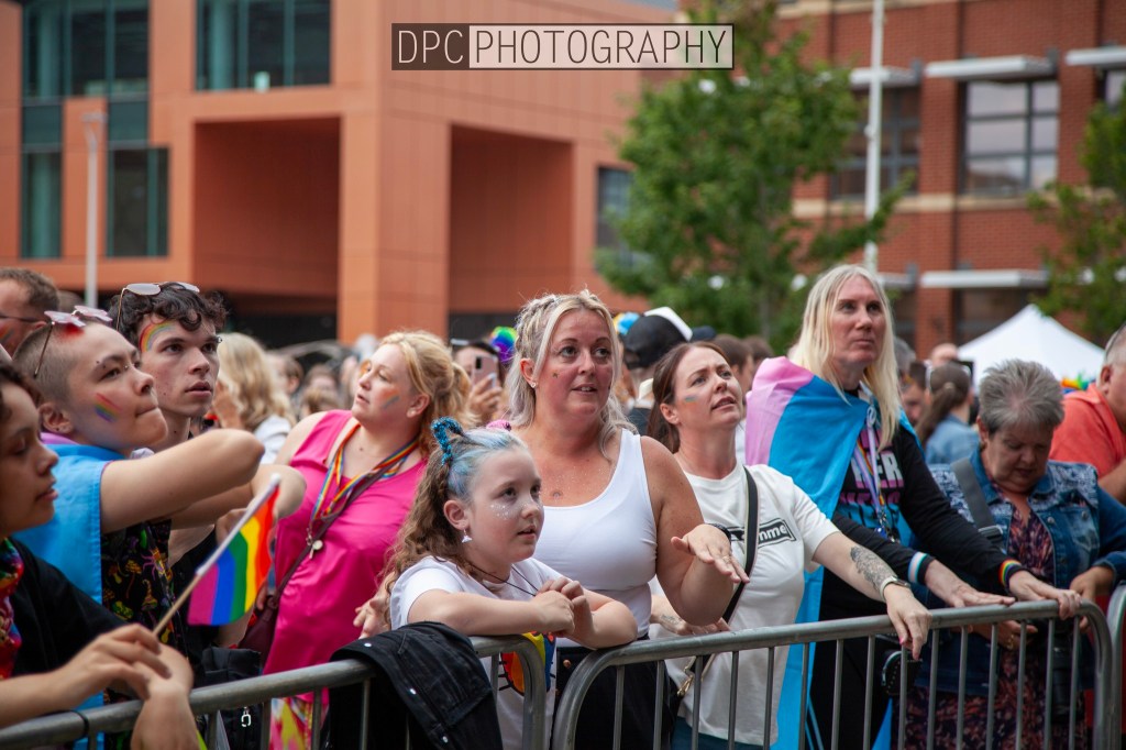 A crowd at a pride event, showing diverse individuals with rainbow flags, colorful clothing, and expressions of excitement and celebration.