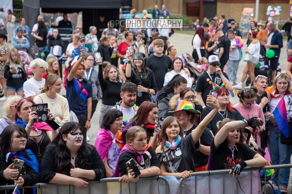 A diverse crowd of people at an outdoor event, many holding phones and wearing colorful attire, including pride flags and accessories, gathered in front of a barrier.