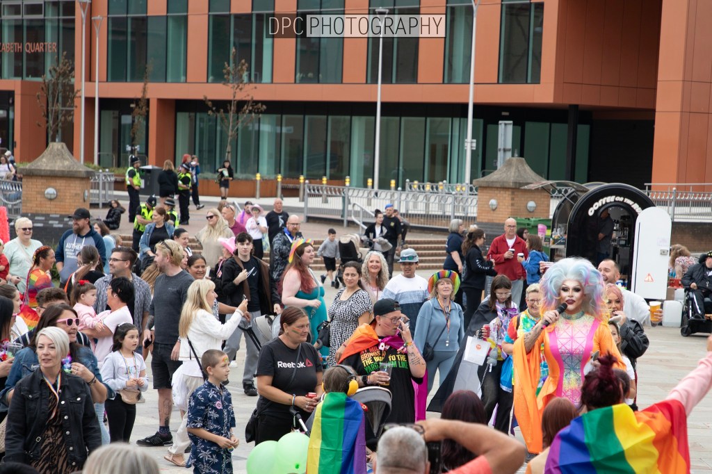 A vibrant LGBTQ+ pride event with a crowd attending, featuring a drag performer on stage wearing a colorful costume and speaking into a microphone, while attendees hold rainbow flags and cheer.
