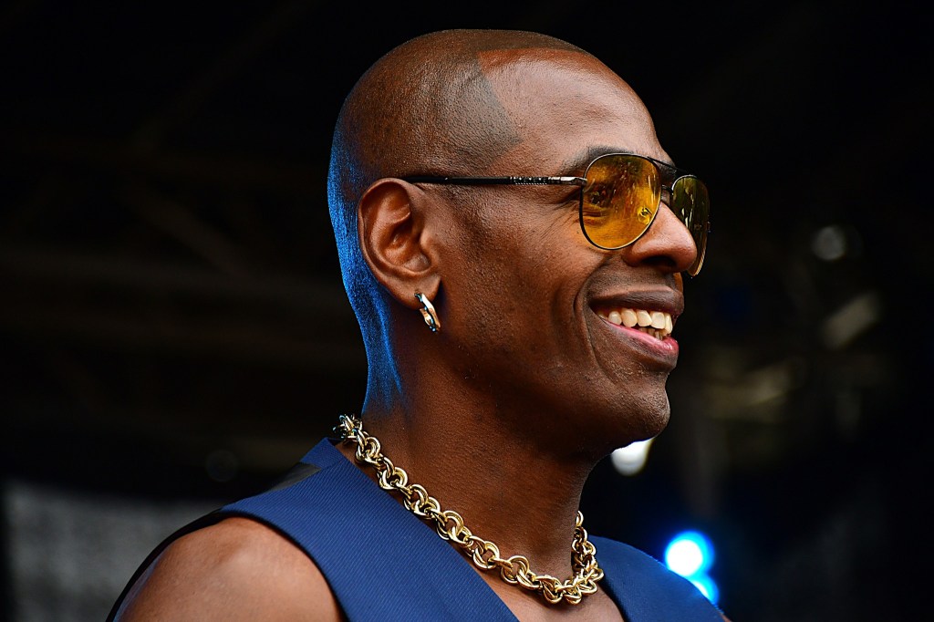 Close-up of a smiling man wearing sunglasses and a chain necklace, with a blurred background.