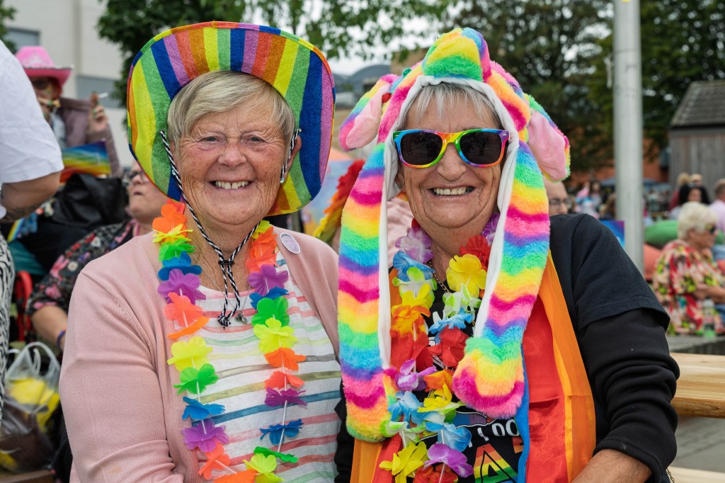 Two smiling women wearing colorful hats and vibrant leis at a festive outdoor event.