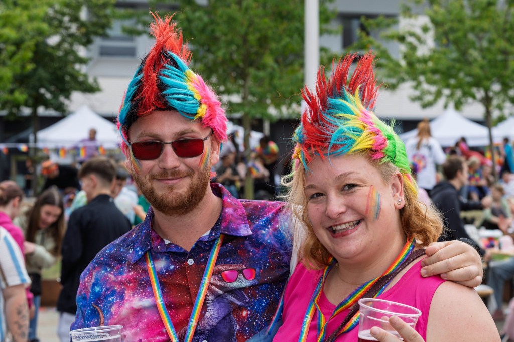 A man and a woman smiling together at a festival, both wearing colorful outfits and vibrant hairstyles. The man has rainbow-colored spiky hair and sunglasses, while the woman has wavy blonde hair with rainbow accents and is wearing a pink top. They are holding drinks and the background features festival attendees and decorations.