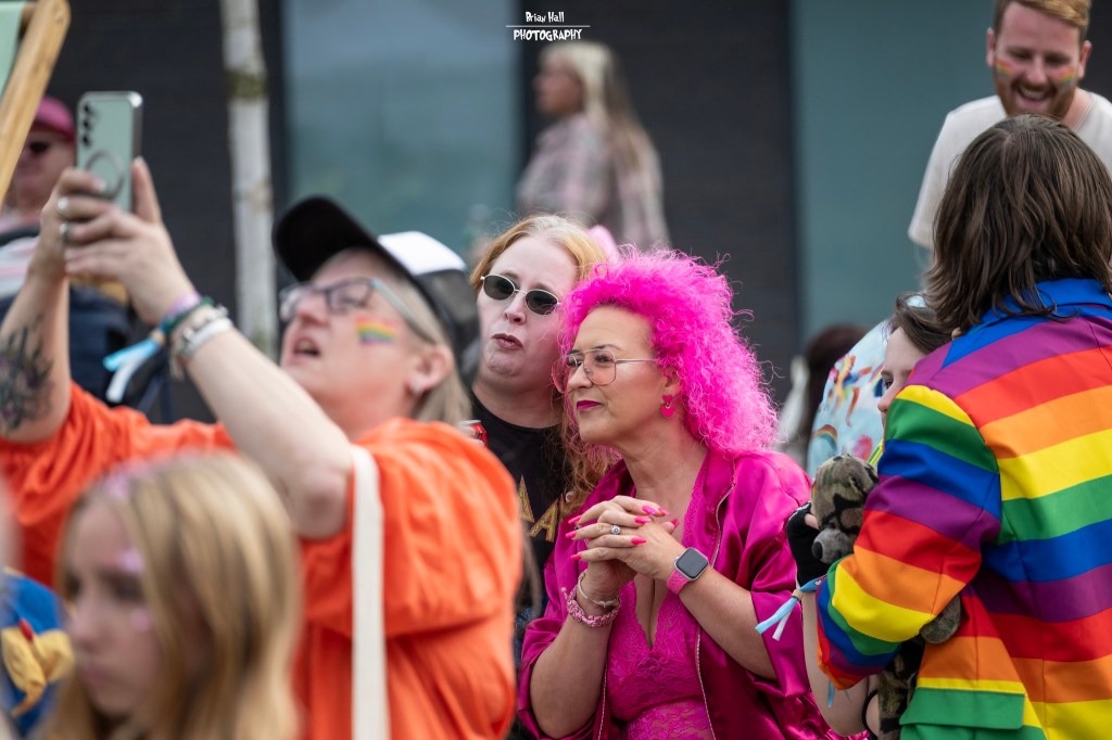 A vibrant crowd at a celebration, featuring a woman with bright pink hair and colorful outfits, capturing the joyful atmosphere with their phones.
