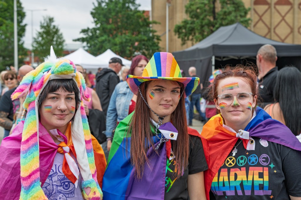 Three individuals celebrating Pride, wearing colorful outfits and accessories, including rainbow flags and a unicorn hat, at an outdoor event.