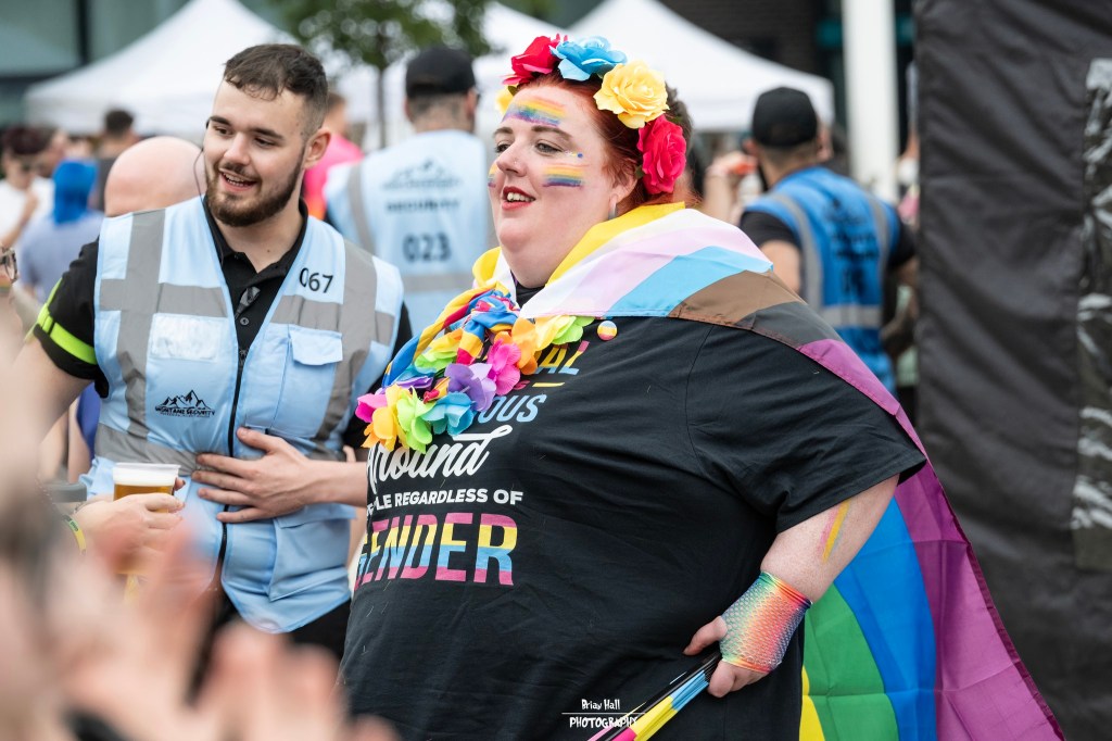 A person with colorful accessories and makeup, wearing a black shirt with a pride message, poses joyfully at an outdoor event, surrounded by spectators and staff in uniforms.