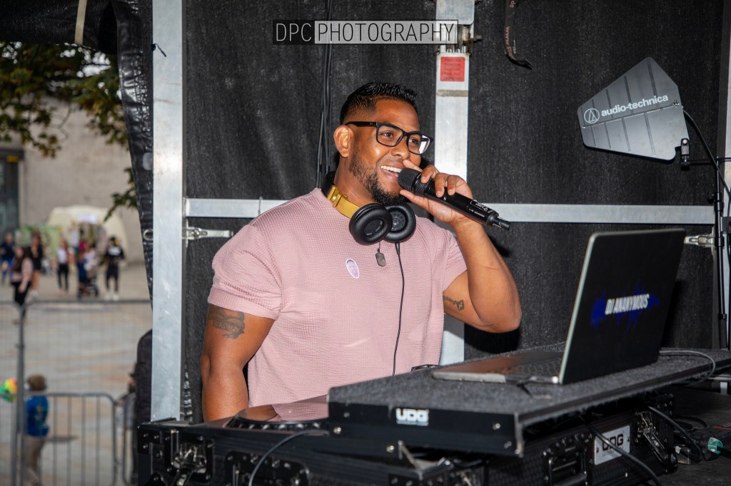 A DJ with glasses and headphones performs at an outdoor event, holding a microphone and interacting with the crowd, while a laptop sits on his DJ equipment.
