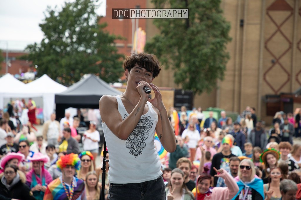 A performer singing on stage in front of a vibrant crowd, with people in colorful outfits and accessories celebrating together at an outdoor event.