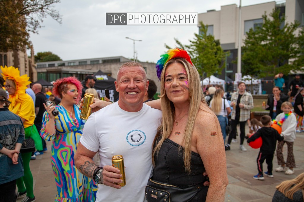 A man and a woman smiling and posing for a photo at a lively outdoor event, surrounded by colorful decorations and attendees in festive outfits.
