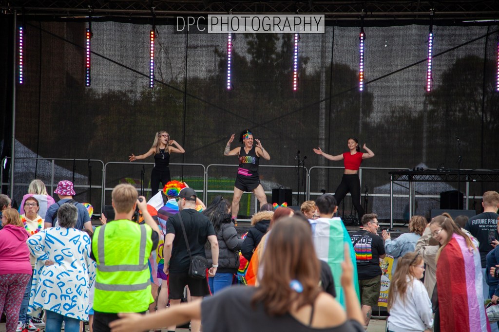 A lively performance at a pride event with dancers on stage and an enthusiastic crowd below, featuring colorful attire and rainbow decorations.