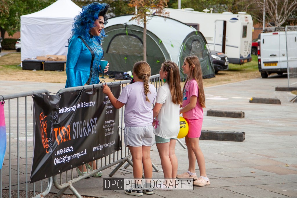 A person in a colorful blue costume and wig interacts with three young girls at a community event, with tents and vehicles visible in the background.