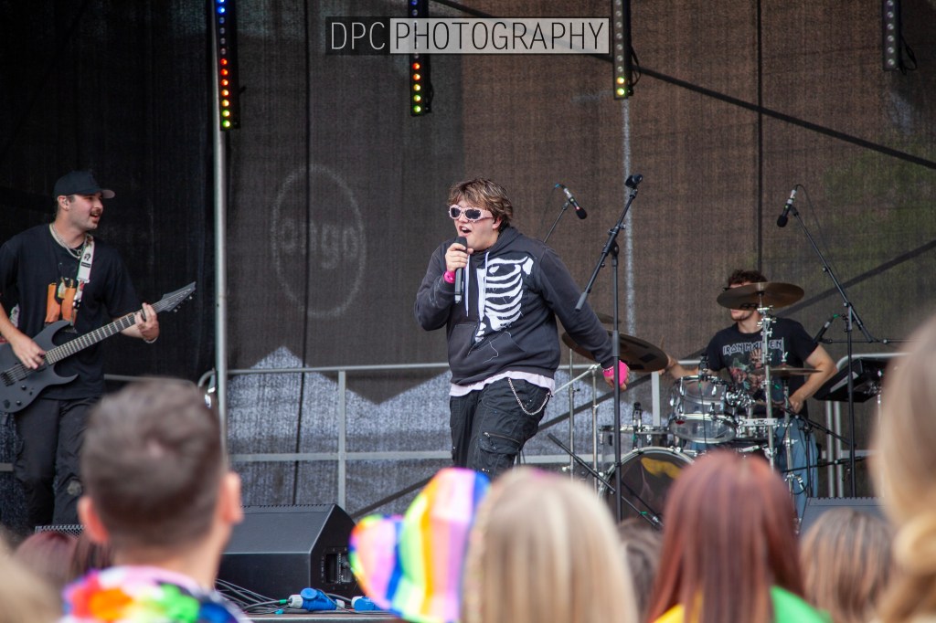 A performer wearing a skeleton sweater sings into a microphone on stage, accompanied by a guitarist and drummer. The audience is visible in the foreground.