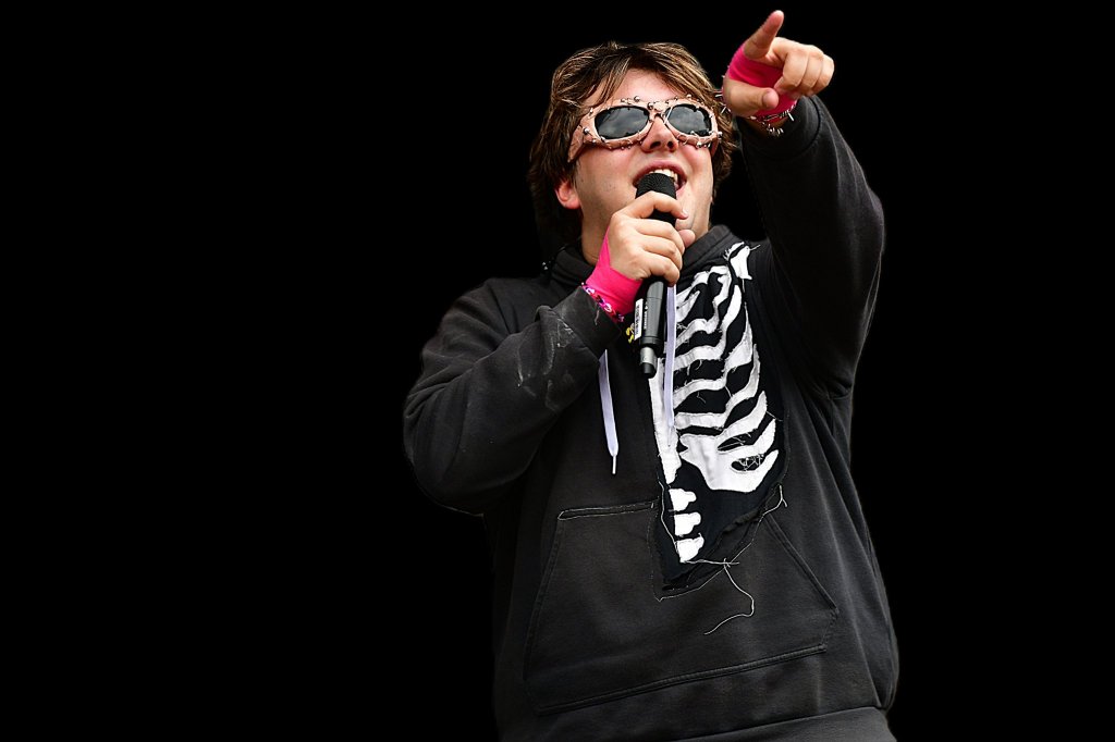 A person wearing sunglasses and a black hoodie with a skeleton design, passionately holding a microphone and pointing while performing.