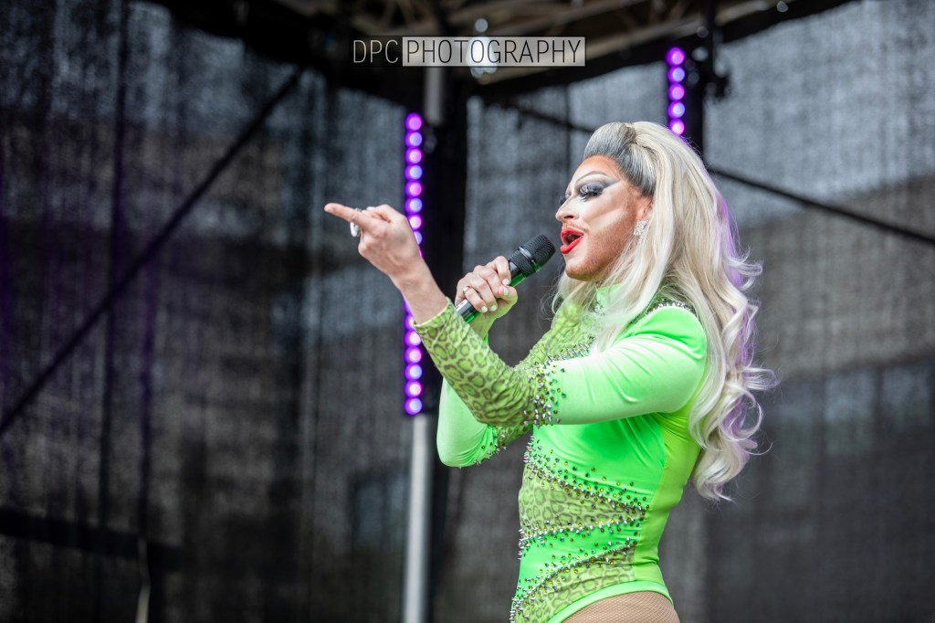 A performer in a vibrant green outfit with rhinestones sings into a microphone on stage.
