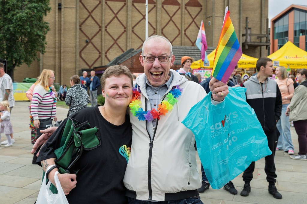 Two smiling individuals posing together at a vibrant outdoor event, one holding a rainbow flag and wearing colorful accessories. Background features event booths and attendees.