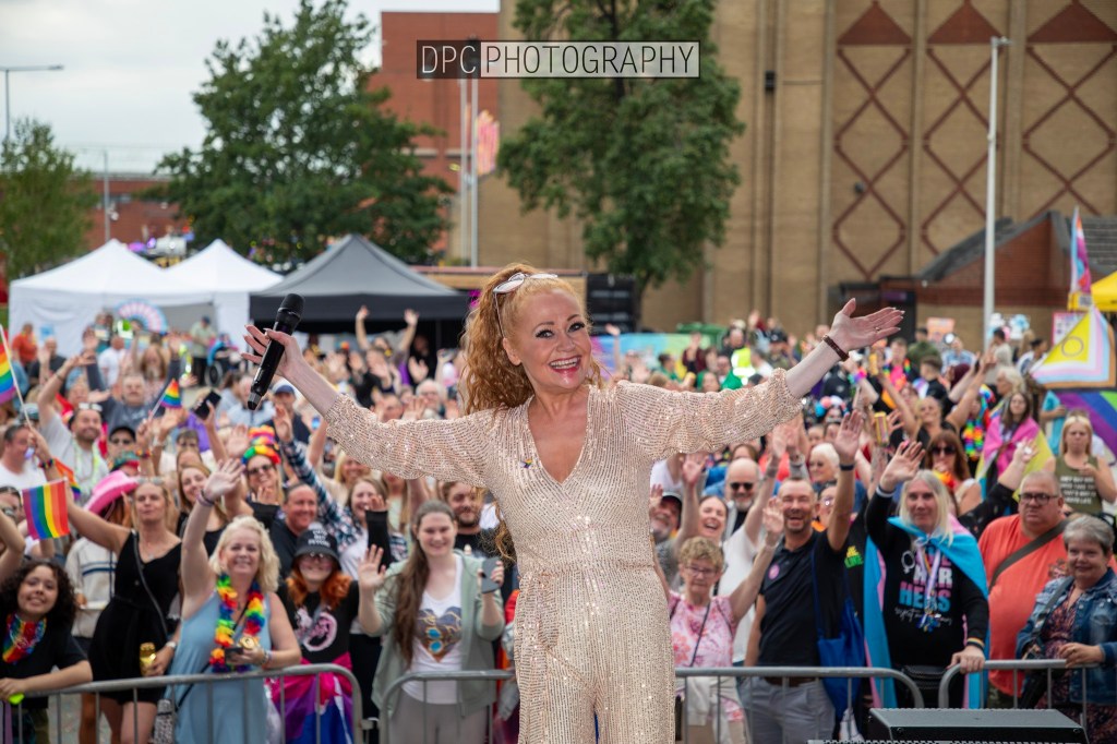 A performer in a sparkling outfit engaging with a lively crowd at a festival, surrounded by people holding rainbow flags and celebrating.