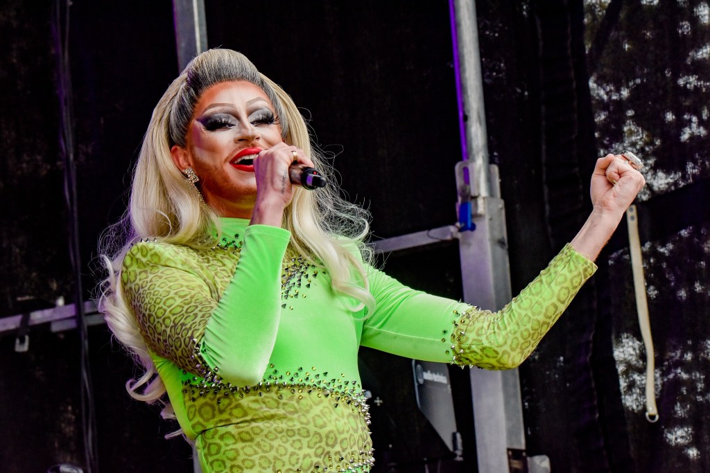 A drag performer with long blonde hair and bold makeup, wearing a bright green outfit with leopard print, passionately singing into a microphone on stage.