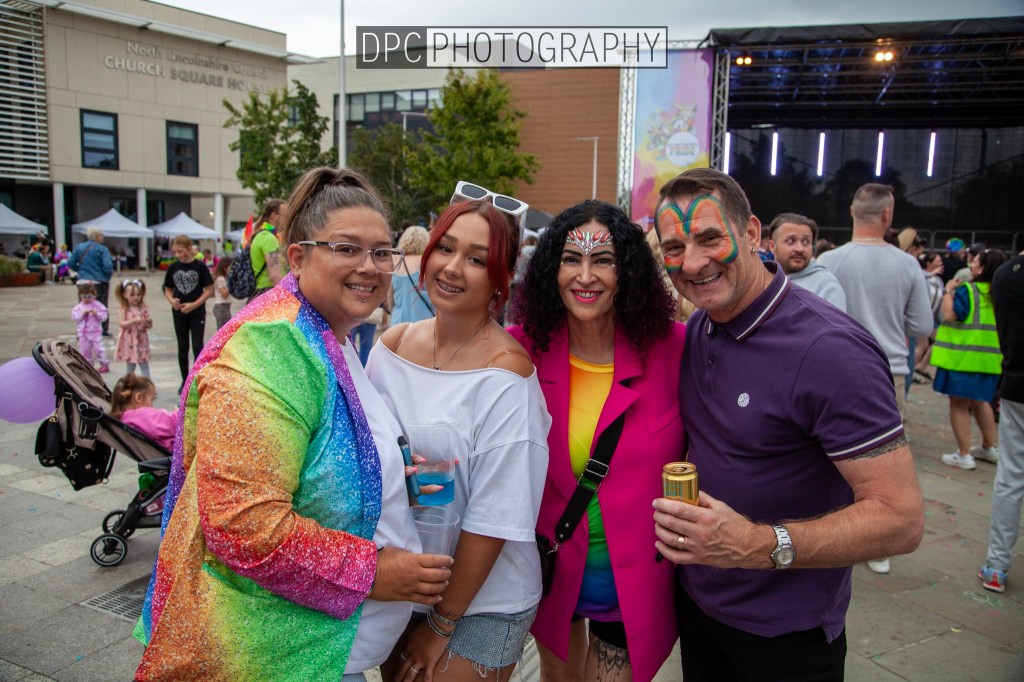 A group of four friends smiling and posing for a photo at an outdoor event, with colorful outfits and festive face paint. The background shows a stage and people enjoying the celebration.