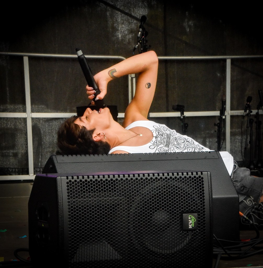 A musician performing passionately on stage while lying on top of a speaker, holding a microphone to their mouth.