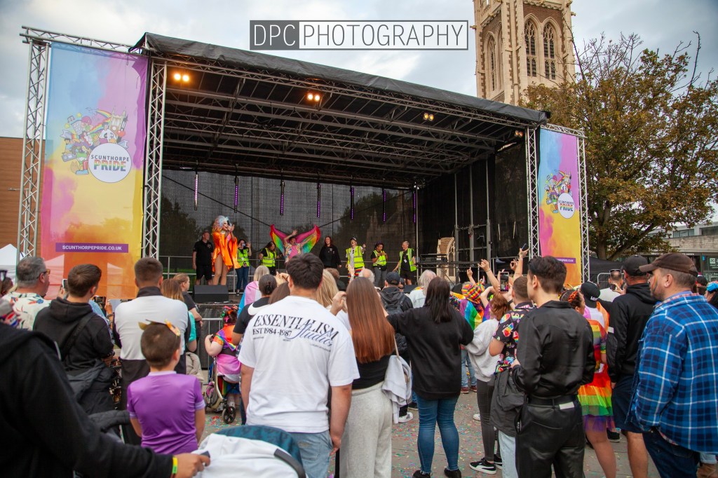 A crowd of people at Scunthorpe Pride, gathered around a stage with a performer in colorful attire, celebrating diversity with rainbow flags.