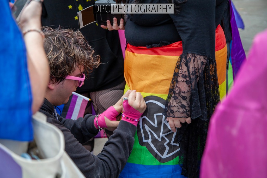 A person wearing pink accessories is decorating a rainbow-colored skirt with a logo at an outdoor event, surrounded by others and colorful flags.
