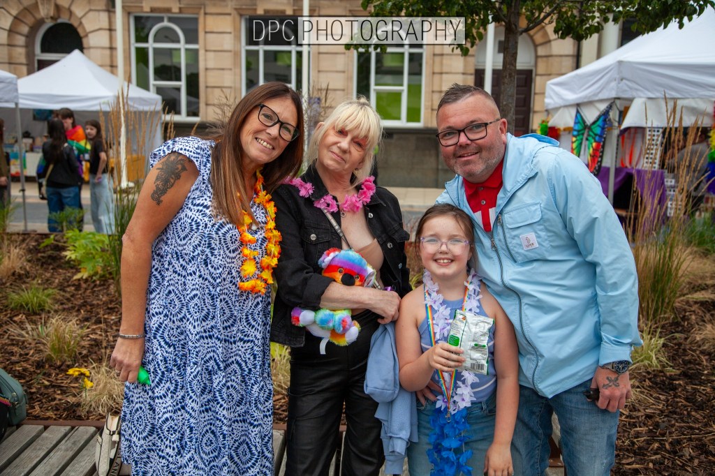 A group of four people smiling and posing for a photo at an outdoor event. They are dressed in colorful attire and accessories, with a festive backdrop of tents and greenery.
