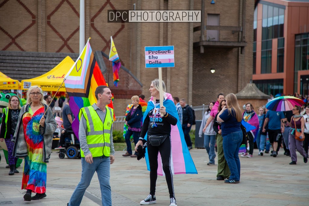 A crowd of people at a event, with someone holding a sign that says 'Protect the Dolls', surrounded by rainbow and trans pride flags.