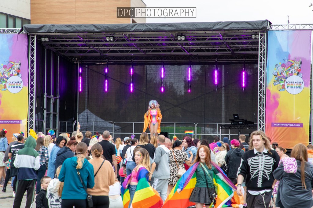 A vibrant crowd at a pride event, gathered in front of a stage with purple lights. A performer in an orange outfit stands on stage, while attendees celebrate with colorful attire and pride flags.