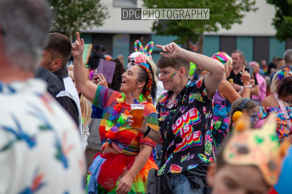 Two people joyfully celebrating in a colorful parade, wearing vibrant outfits and accessories, while others are visible in the background.