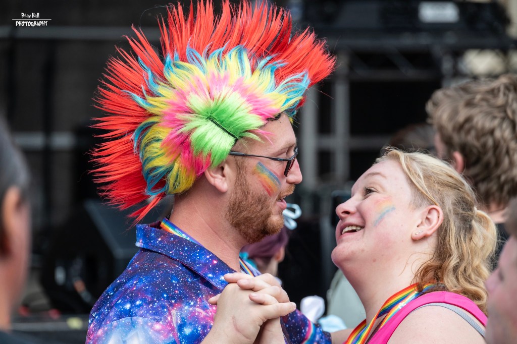 A couple smiling at each other while dancing, with one person wearing a colorful spiked wig and vibrant attire, set against a festival backdrop.