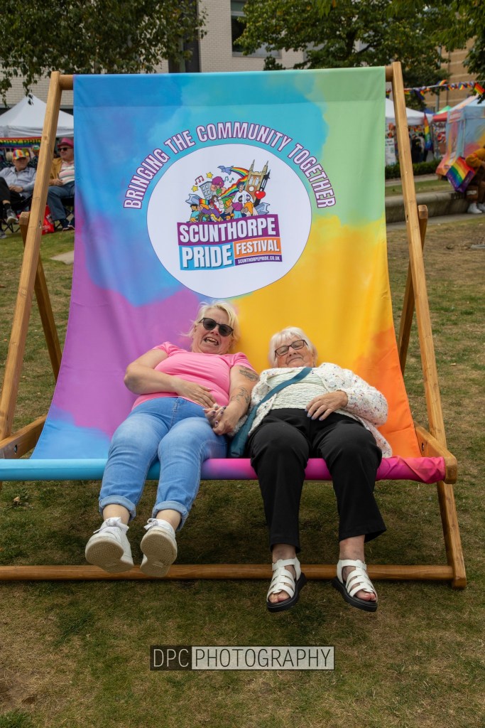 Two women enjoying a colorful lounge chair at the Scunthorpe Pride Festival, smiling and relaxing together.