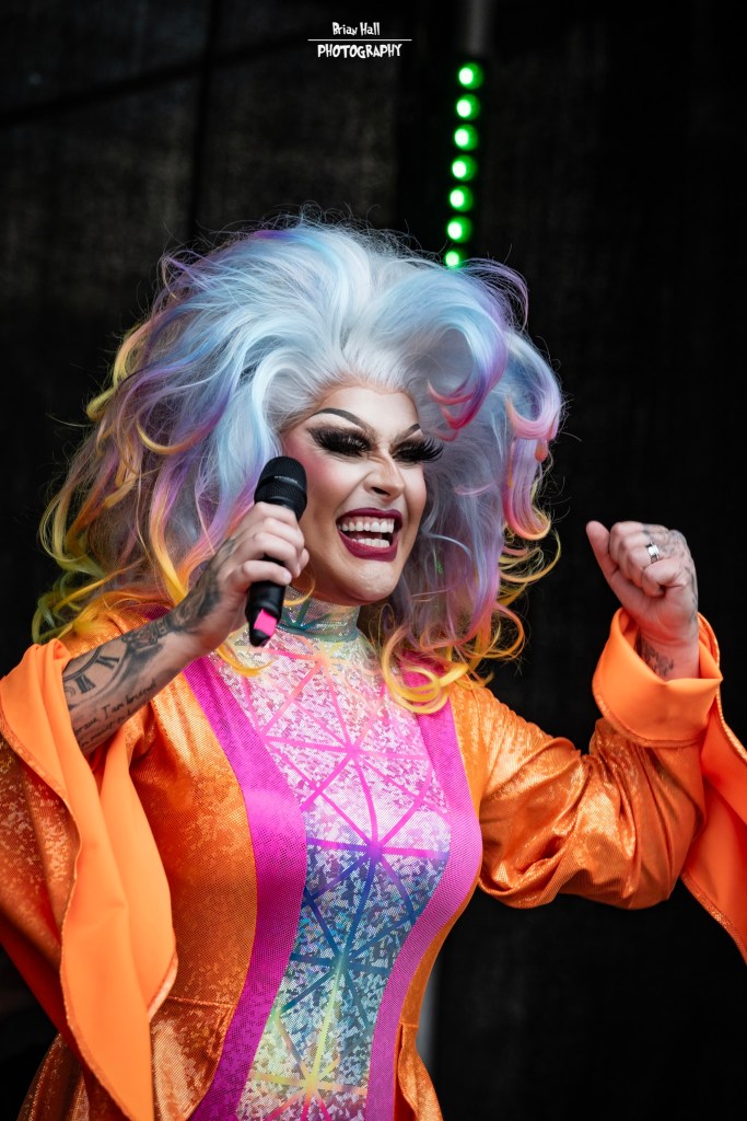 A performer with vibrant, multi-colored hair and a striking orange costume passionately sings into a microphone on stage.