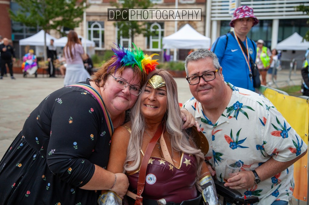 A group of three people posing together, one wearing a superhero costume, with colorful hairstyles and clothing. In the background, tents and trees are visible.